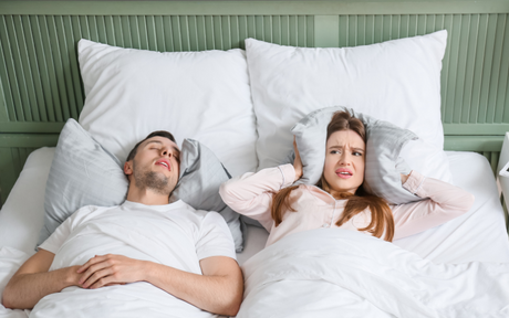 Woman covering ears with pillow while man sleeping next to her snores.