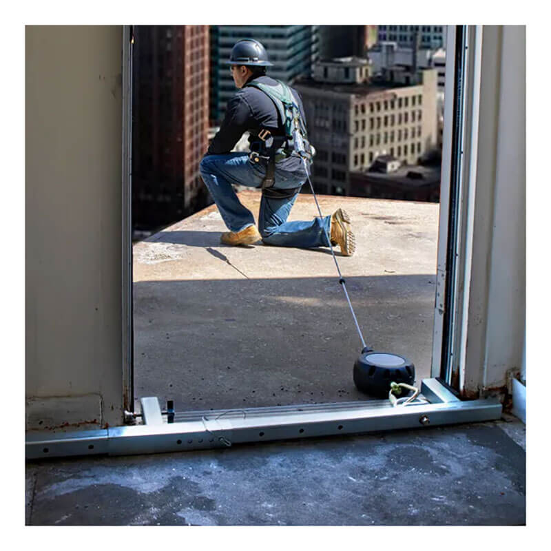 A worker in safety gear kneels at a rooftop edge, secured by a French Creek Production Door Jamb Anchor 1785 harness for OSHA-compliant fall protection. City buildings appear in the background.