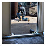 A worker in safety gear kneels at a rooftop edge, secured by a French Creek Production Door Jamb Anchor 1785 harness for OSHA-compliant fall protection. City buildings appear in the background.